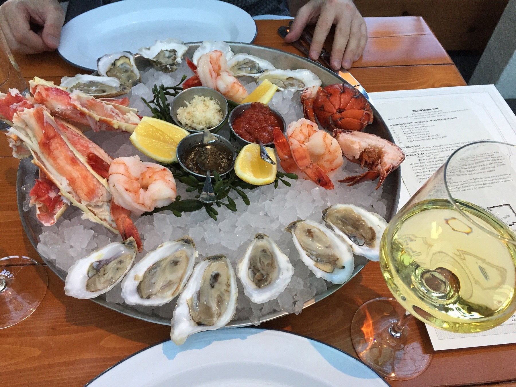 A chilled seafood platter on a bed of ice featuring raw oysters, crab legs, shrimp, lobster tail, and lemon wedges, with dipping sauces in the center and a glass of white wine nearby.