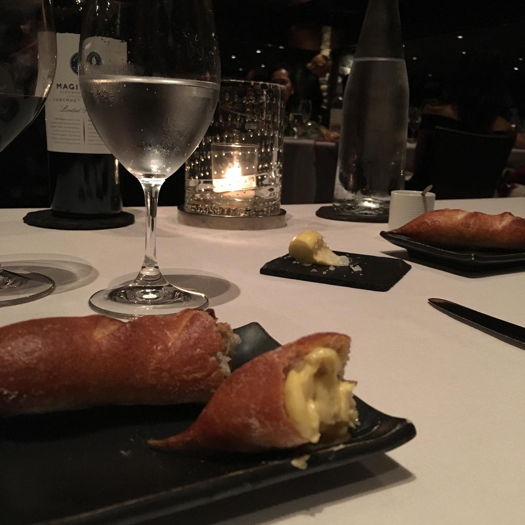 Two small bread rolls served on dark plates at bar seating, one sliced and spread with butter, next to wine and water glasses on a white tablecloth.