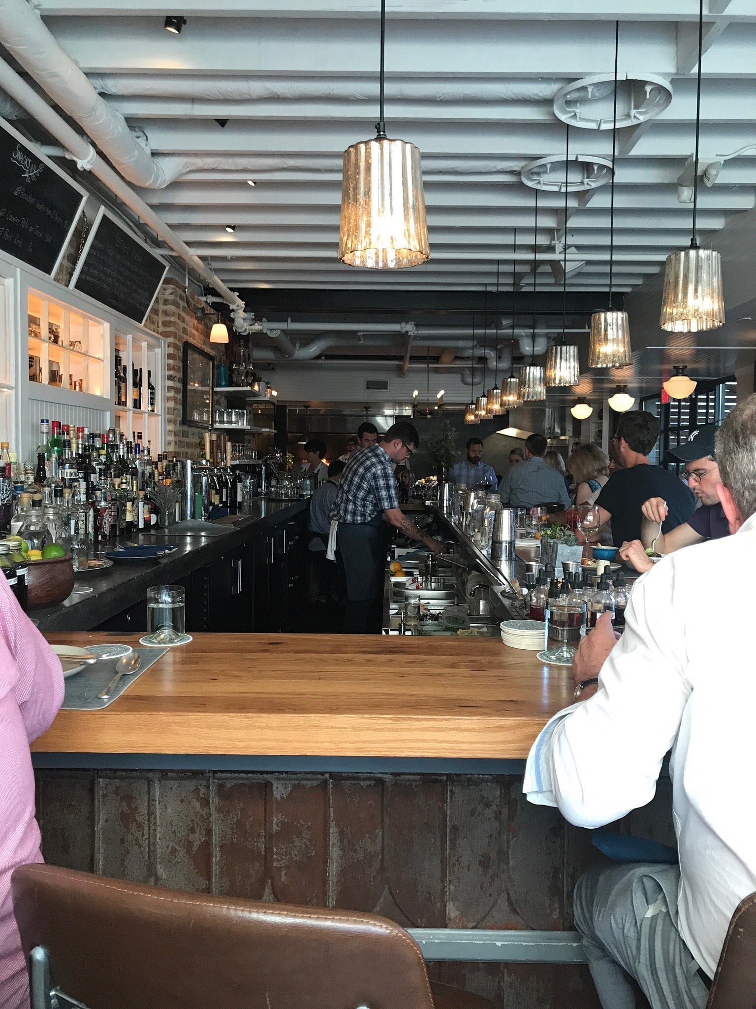 Customers sit at long wooden bar seating as a bartender prepares drinks.