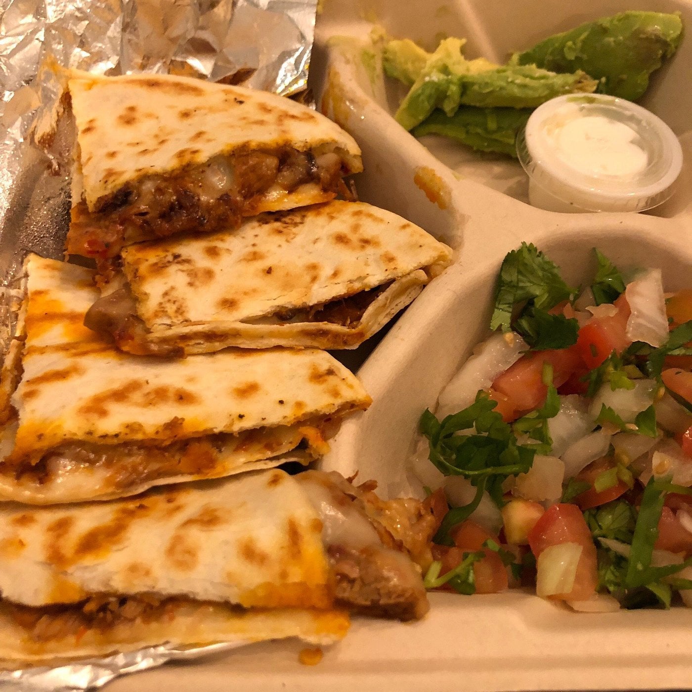 A take-out container with a quesadilla cut into triangles, a side of avocado slices, a small cup of sour cream, and a serving of pico de gallo.