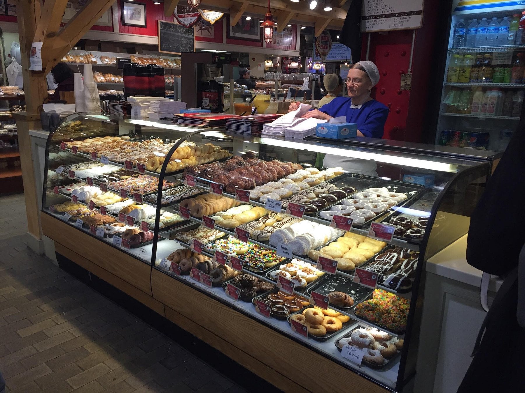 A baker behind a glass display case filled with a variety of donuts and baked goods at a market stall.
