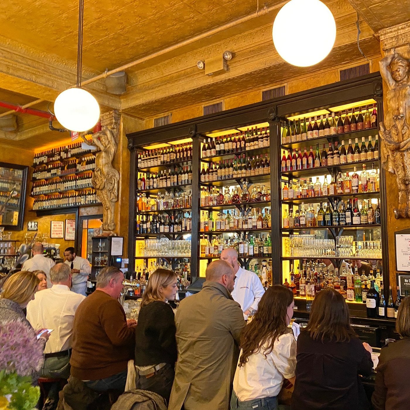 Patrons enjoy bar seating at a counter beneath a tall, multi-tiered shelf stocked with a large collection of wine and liquor bottles.