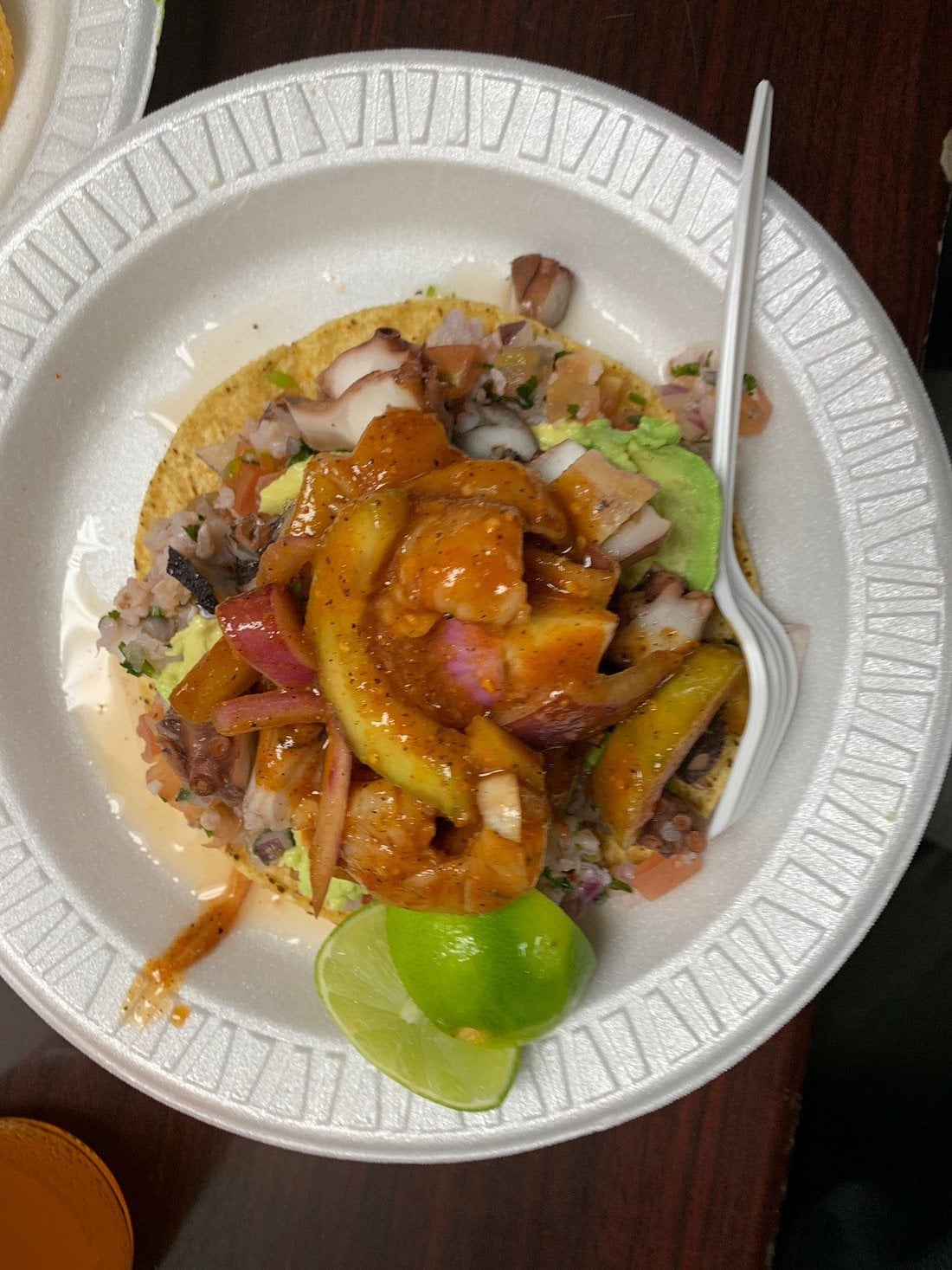A seafood tostada topped with shrimp, octopus, and avocado on a styrofoam plate, served with a lime wedge and a plastic fork.