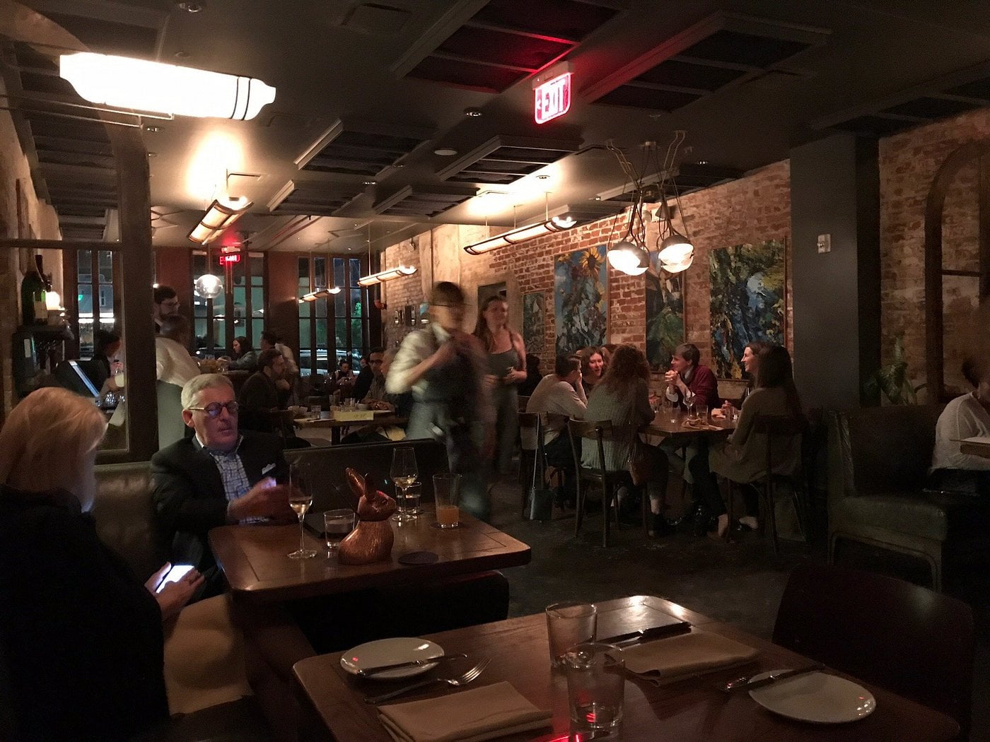 Dimly lit restaurant dining room with patrons sitting at wooden tables and a staff member walking through the center aisle.