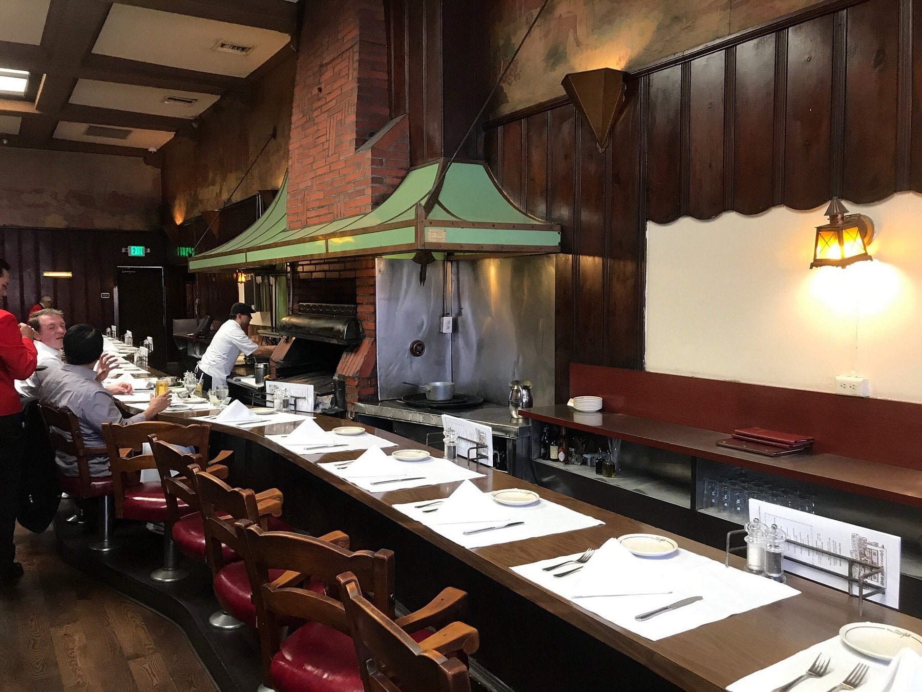 A chef cooks at a brick grill behind a curved restaurant counter with bar seating and place settings, where a few patrons are seated.