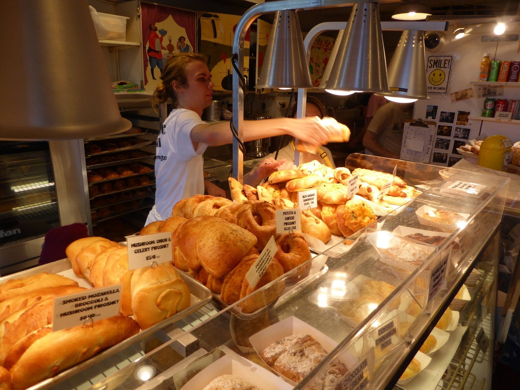 A food counter employee reaches for a fresh pastry behind a glass display case filled with rows of labeled bread and savory piroshki.