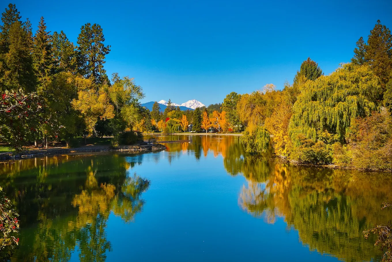 A calm lake reflects autumn trees and a clear blue sky, with a snow-capped mountain in the background. The scene is bright and peaceful, surrounded by colorful foliage and tall evergreens. Once-affordable retirement towns.