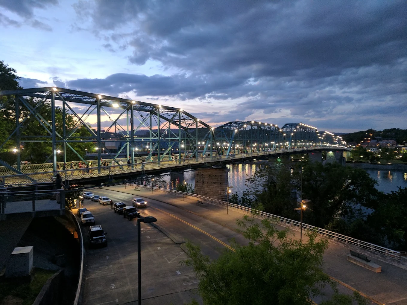 A lit steel truss bridge crosses over a river at dusk. Cars and people are on the bridge, with streetlights reflecting on the water. The sky is cloudy with hints of sunset colors.