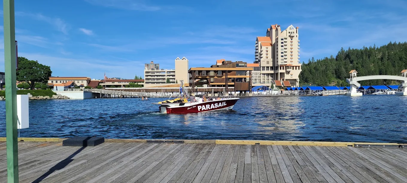 A red parasailing boat is moving on a lake near a dock, with waterfront buildings and a forested hillside in the background under a clear blue sky.