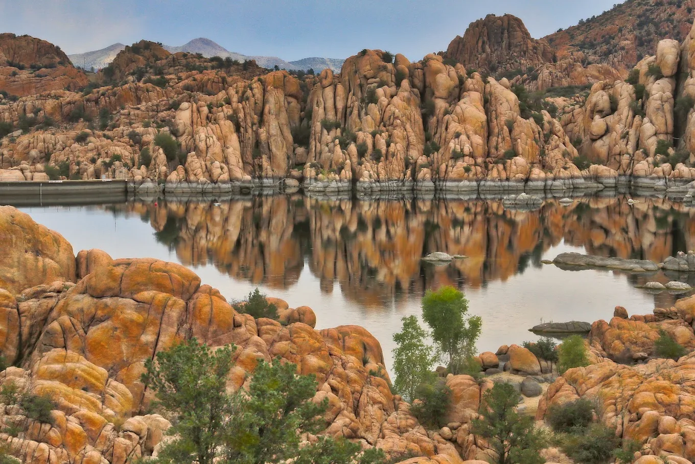 Orange and tan rocky cliffs reflect in a calm lake, surrounded by sparse green vegetation and distant mountains under a cloudy sky.