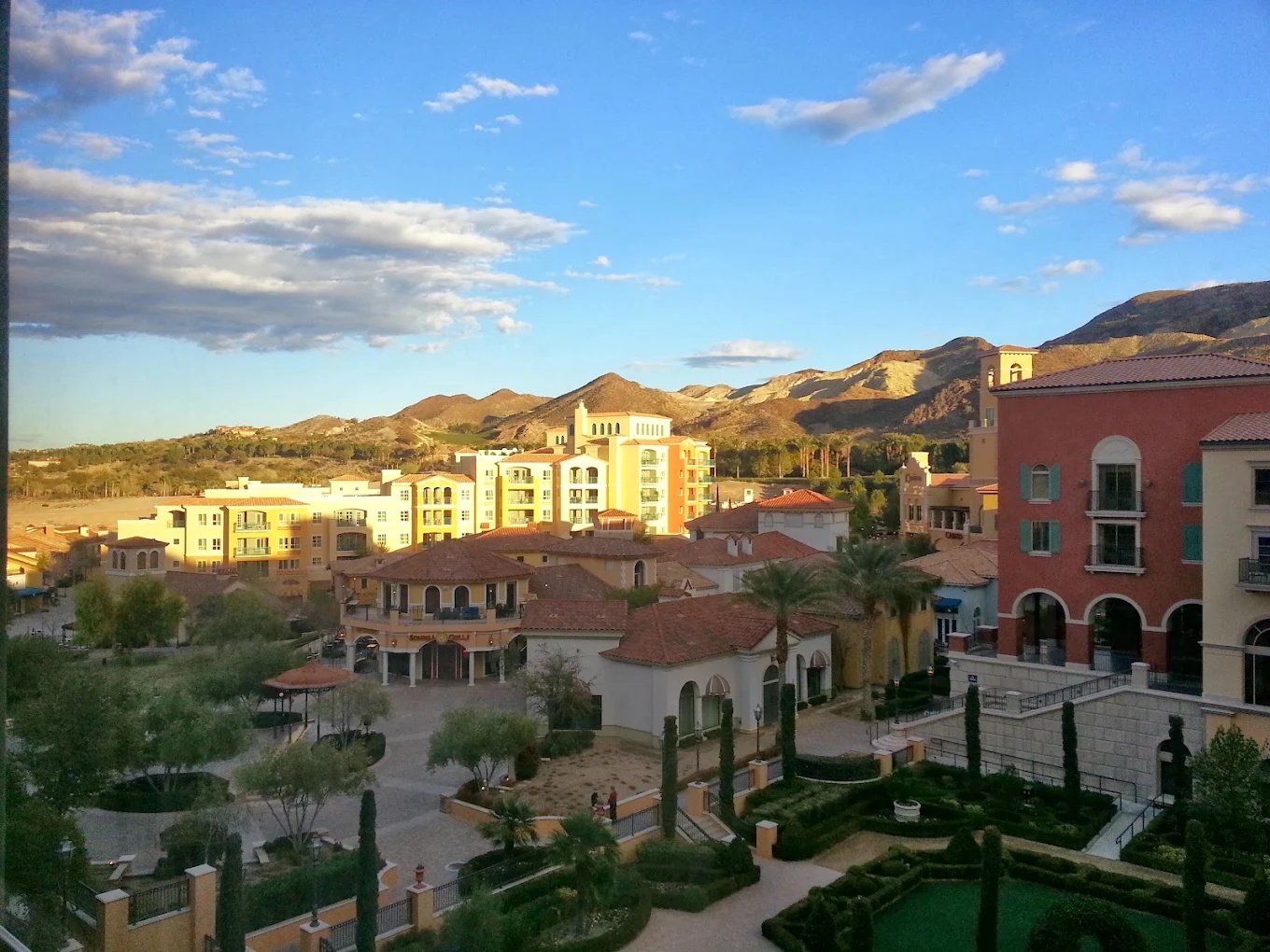 A scenic view of Mediterranean-style buildings with red-tiled roofs, surrounded by gardens and palm trees, set against mountains under a blue sky with scattered clouds.