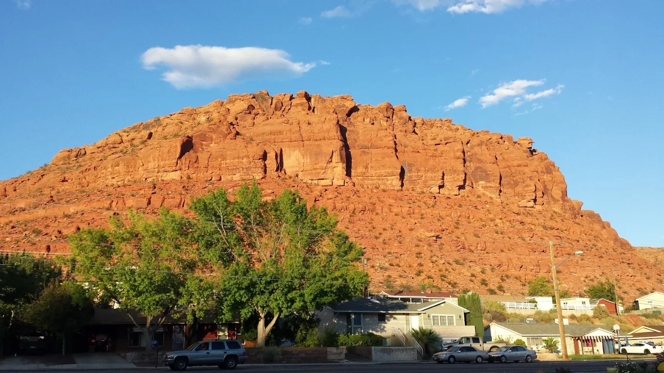 A large red rock mesa rises behind green trees and houses under a blue sky with scattered clouds. Cars are parked along the street in front of the homes.