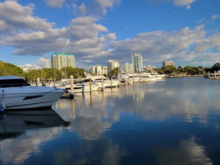 Yachts docked at a marina with calm water reflecting boats, sky, and clouds; tall buildings and trees in the background under a partly cloudy sky.