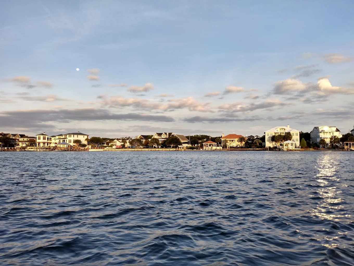 Calm water with gentle ripples in the foreground, a shoreline lined with houses and trees in the background, and a blue sky with scattered clouds above.