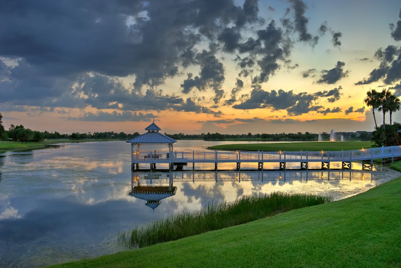 A wooden pier with a gazebo extends over a calm lake at sunset, reflecting clouds and colorful sky. Green grass and palm trees line the shore.