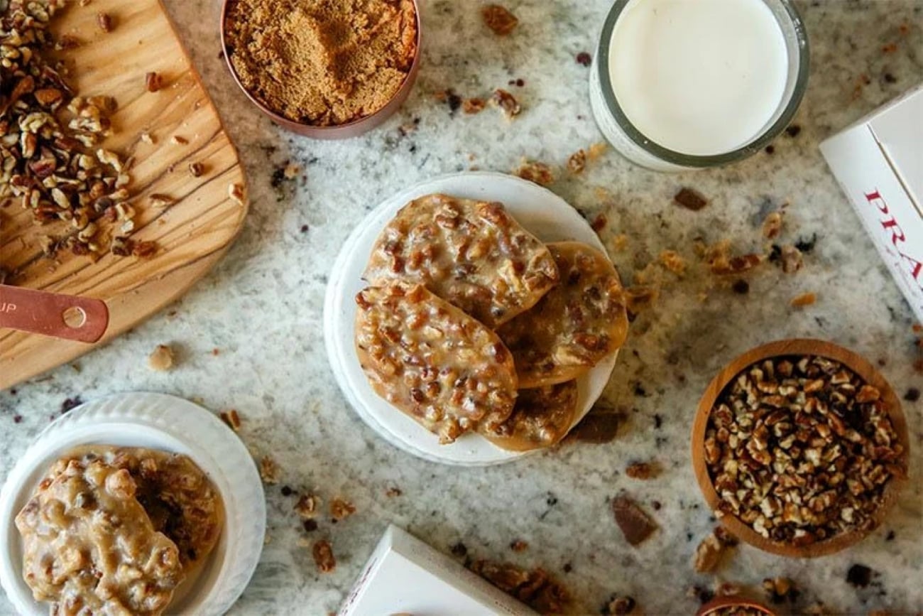 A stack of the best souvenir pecan pralines on a white plate, surrounded by bowls of pecans, brown sugar, and a glass of milk on a marble countertop.