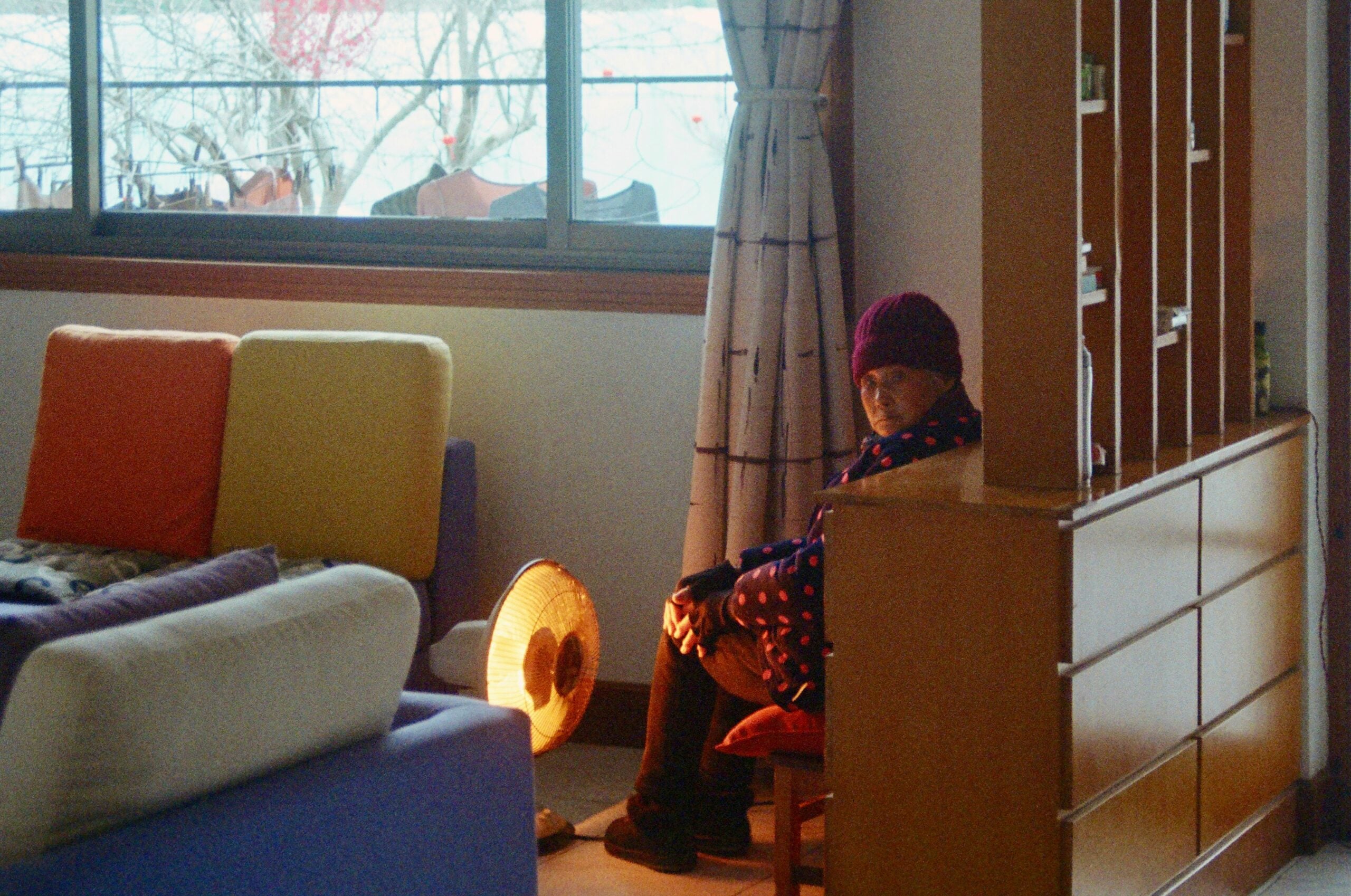 An elderly person in a red hat and polka dot coat sits on a chair indoors beside a heater, with sunlight coming through a window and colorful couches nearby.