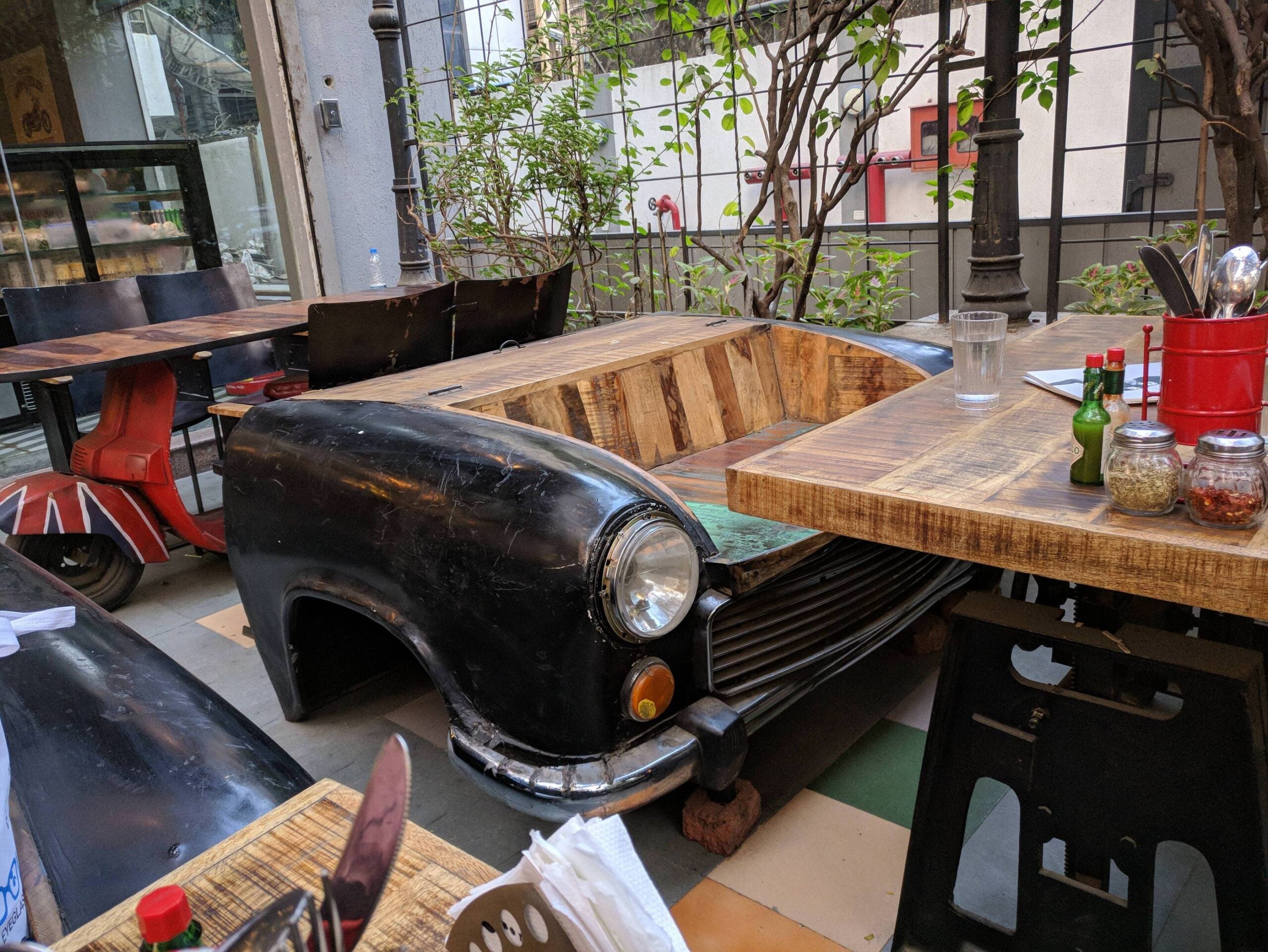 A restaurant seating area uses a vintage car body as a bench at a wooden table, surrounded by chairs and plants, creating a quirky and retro dining atmosphere.