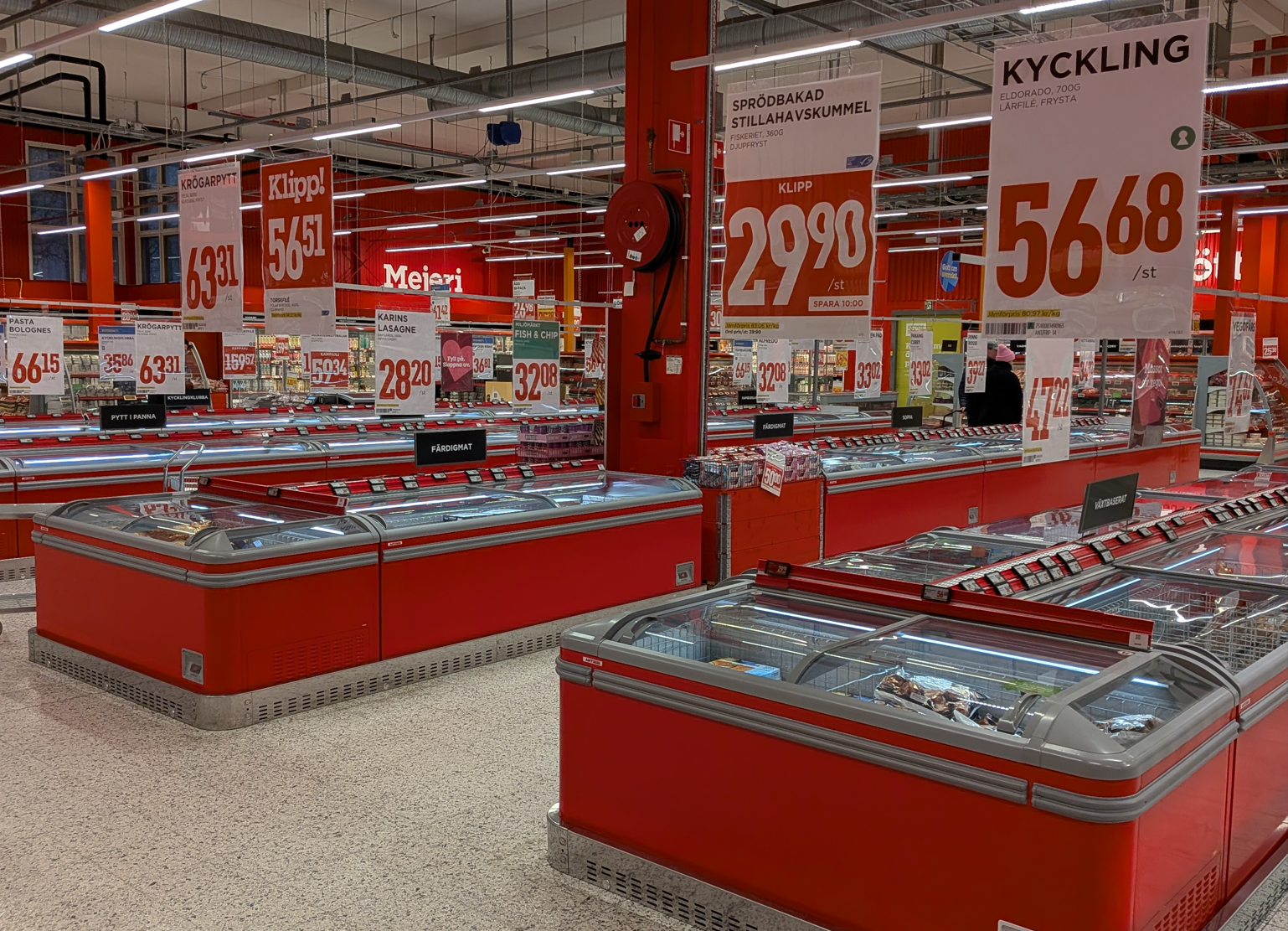 Rows of red refrigerated display cases in a supermarket with hanging price signs for various frozen food products.