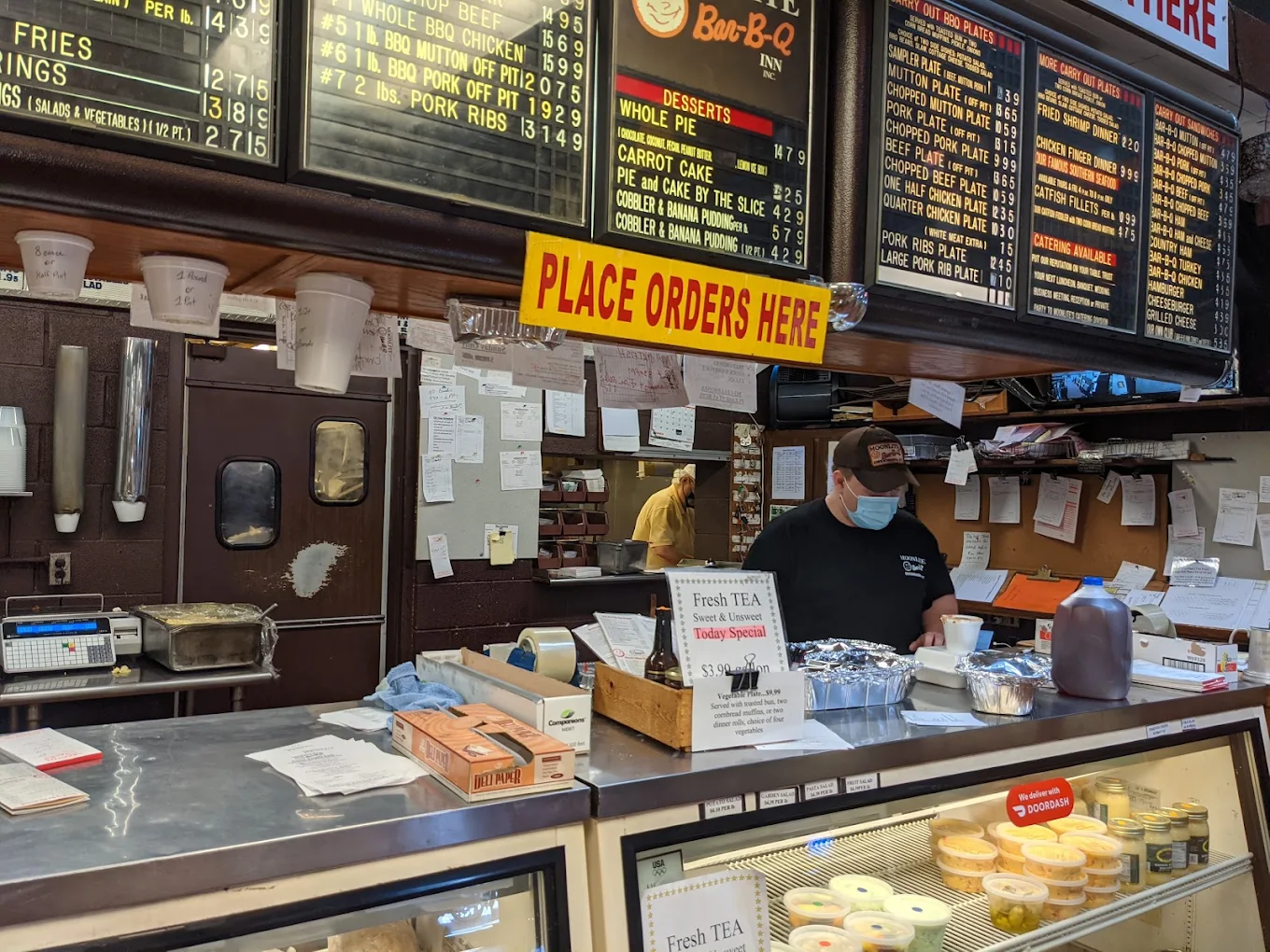 A masked employee behind a restaurant counter works below large menu boards and a yellow “Place Orders Here” sign. Forgotten American foods.