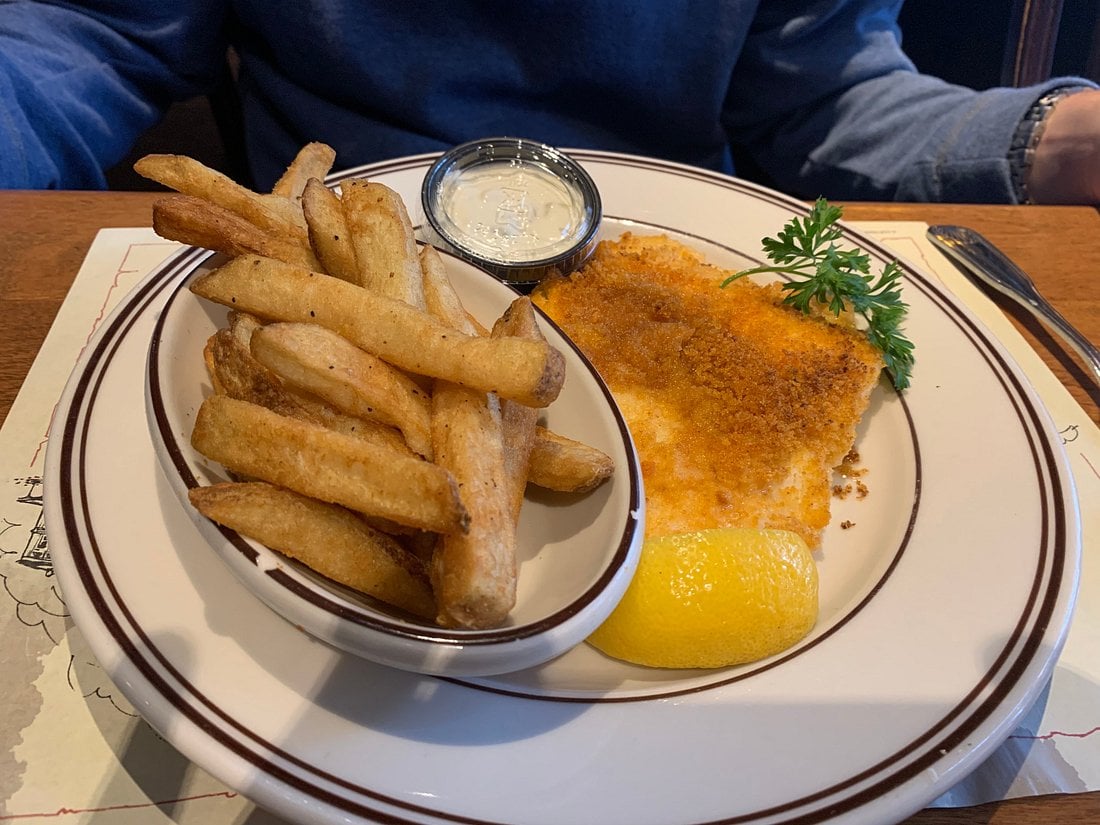 A plate featuring a breaded fish fillet, a lemon wedge, a sprig of parsley, and a side dish of french fries with a small container of dipping sauce.