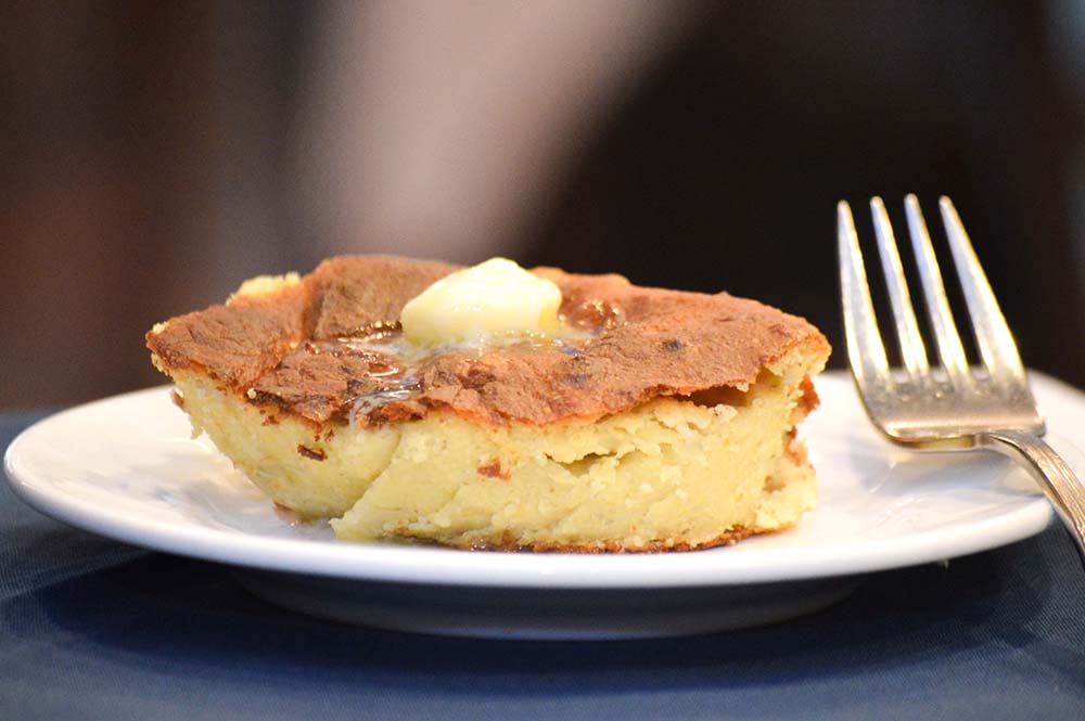 A slice of baked potato cake topped with a melting pat of butter, served on a white plate with a fork.