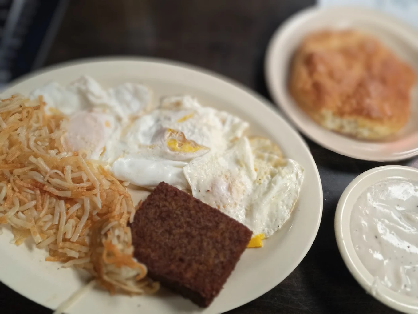 A breakfast plate with fried eggs, shredded hash browns, and a square of dark toast, accompanied by a small bowl of gravy and a separate biscuit.