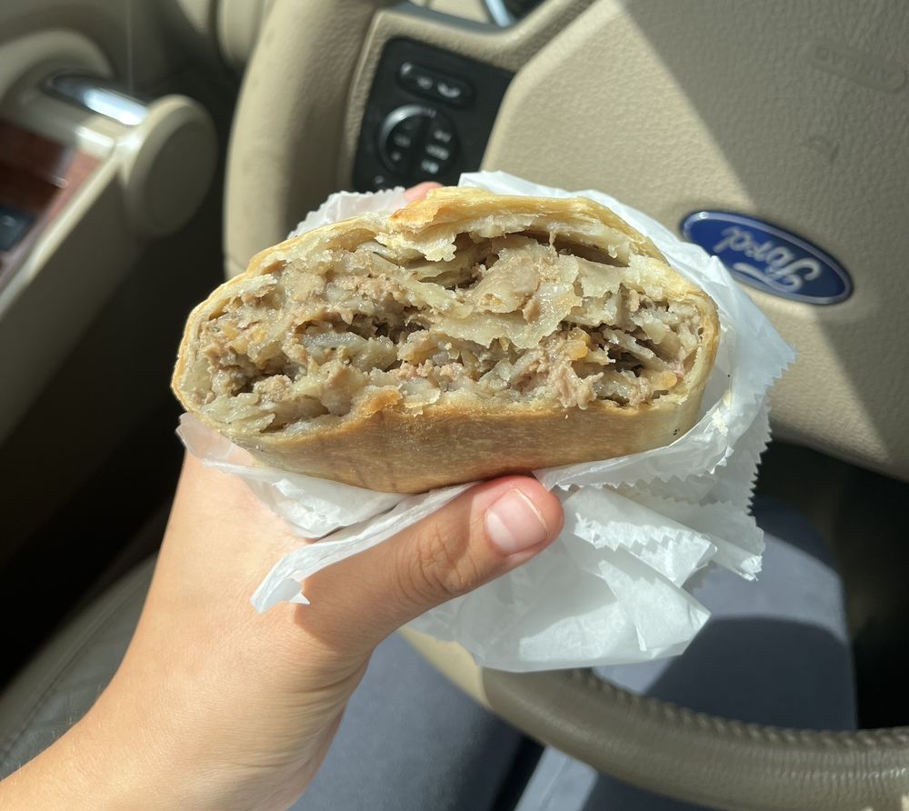 A hand holding a partially eaten savory meat and potato pastry wrapped in white paper inside a car.