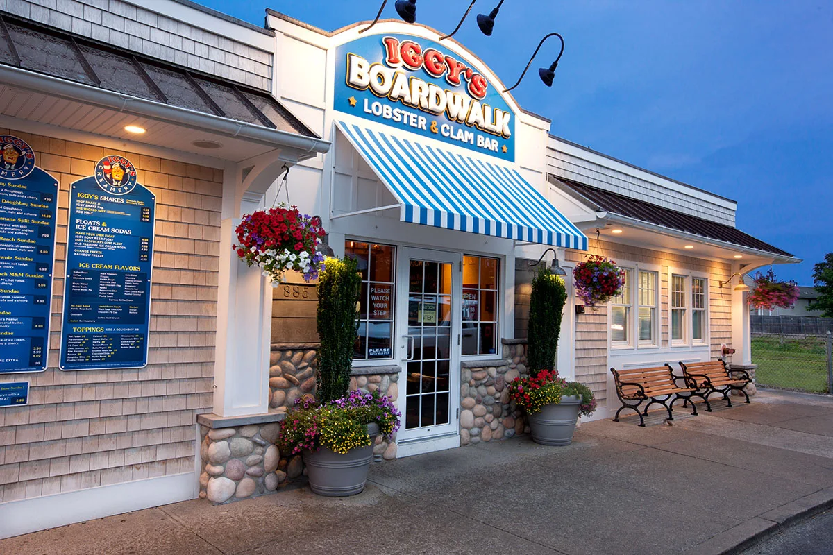 Iggy’s Boardwalk Lobster and Clam Bar restaurant entrance with a menu board on the wall, blue and white striped awning, and two wooden benches outside.