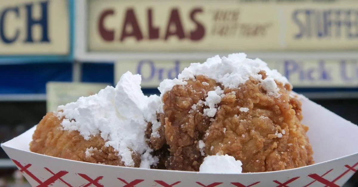 Two fried calas fritters topped with powdered sugar in a paper tray in front of a food stand sign.