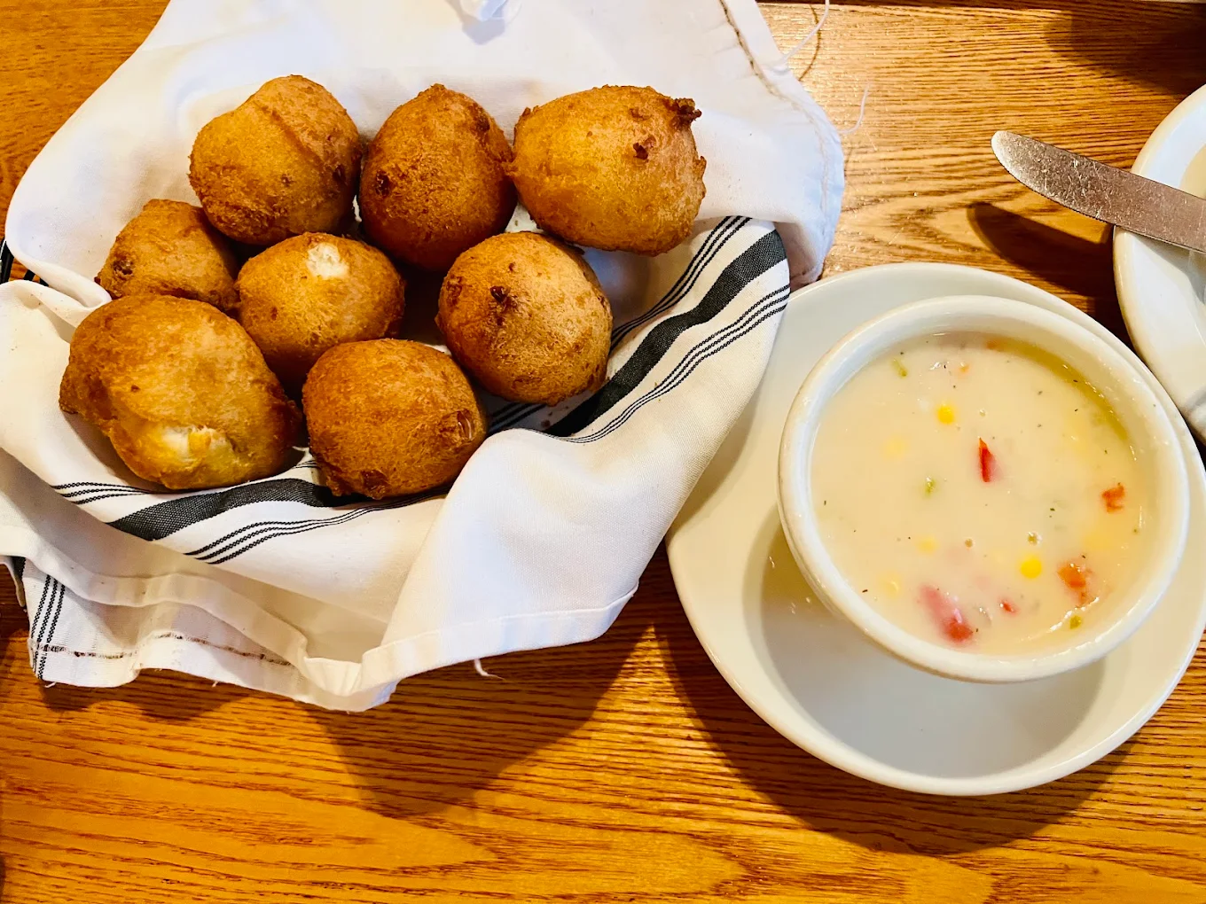 A basket of fried hushpuppies next to a bowl of creamy corn chowder on a wooden table.