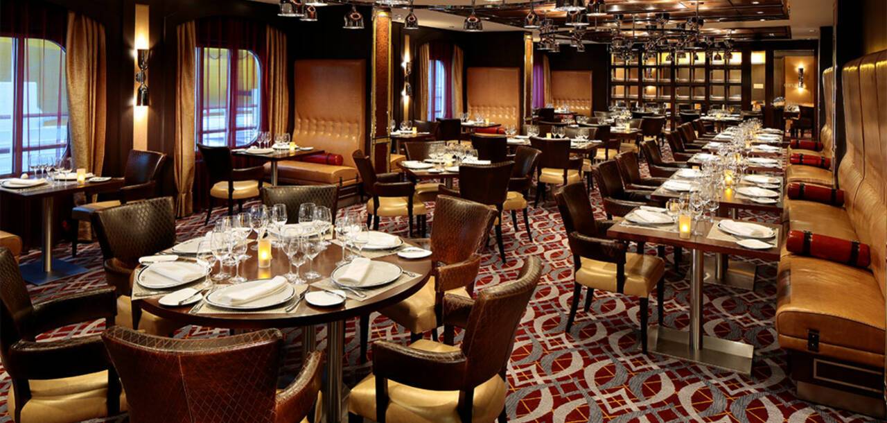 Dining room of a restaurant with round and rectangular tables set with glassware and plates, surrounded by upholstered chairs and leather banquettes.