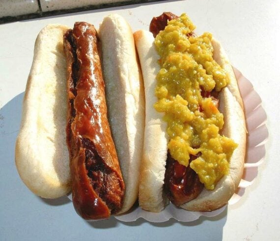 Two hot dogs in buns on a paper plate, one plain with glaze and the other topped with yellow relish.