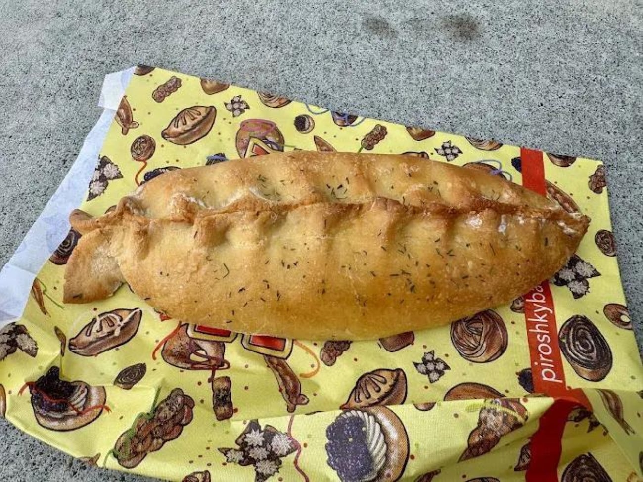 A golden-brown baked piroshky sprinkled with herbs, resting on a patterned yellow paper bag.