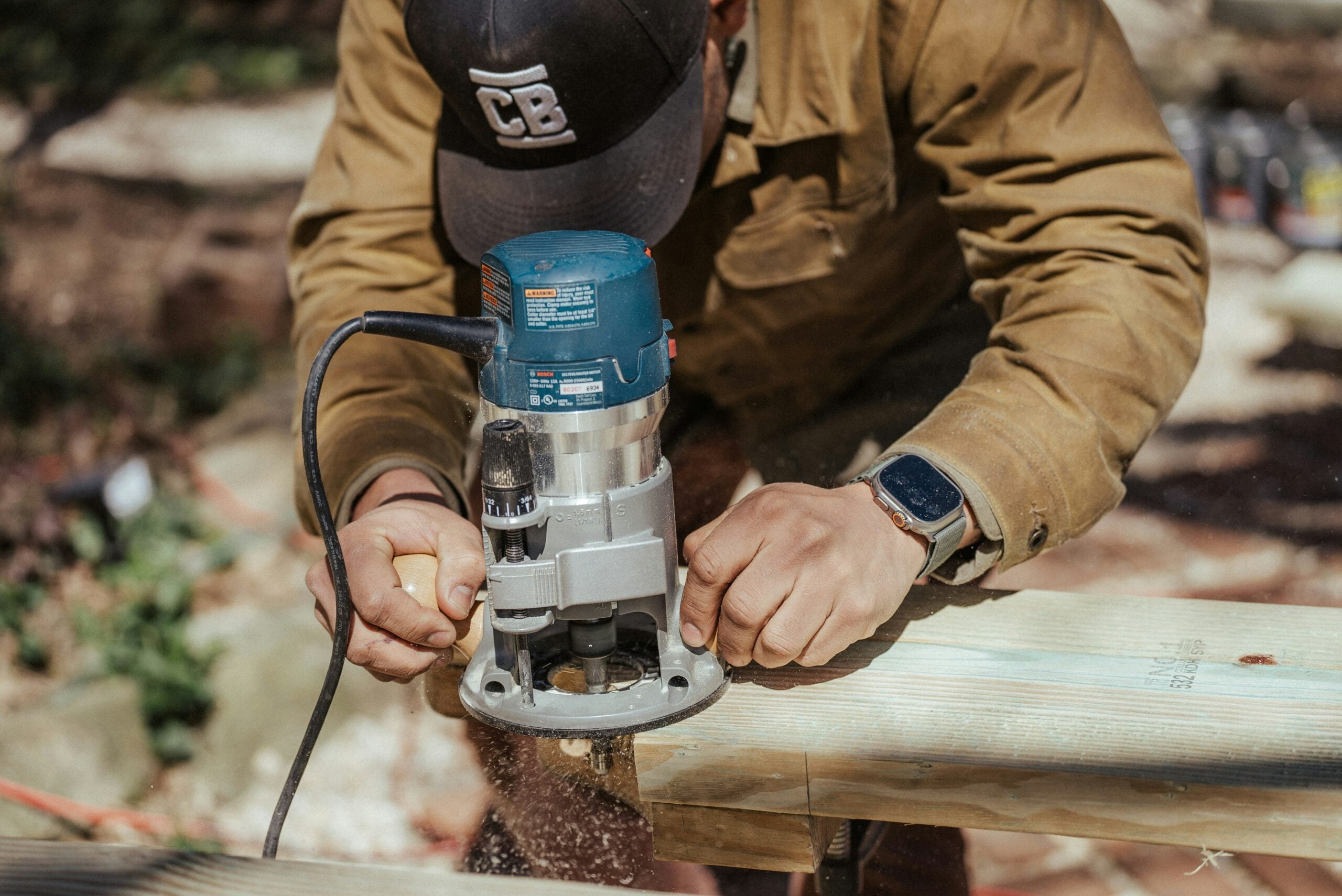 A person in a brown jacket and baseball cap uses a blue power router to shape the edge of a wooden board.