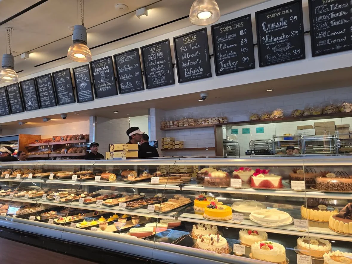 A long refrigerated display case filled with cakes and pastries in a bakery, with menu boards listing food items on the wall behind the counter. Best bakeries for lunch.