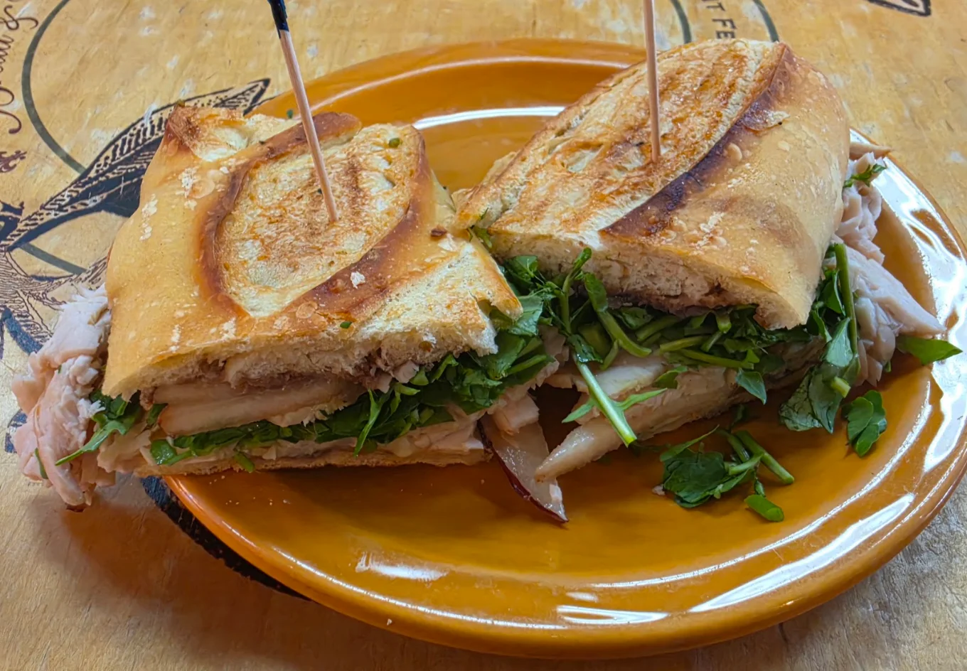 A turkey sandwich on a baguette with greens, held together by two toothpicks on an orange plate.