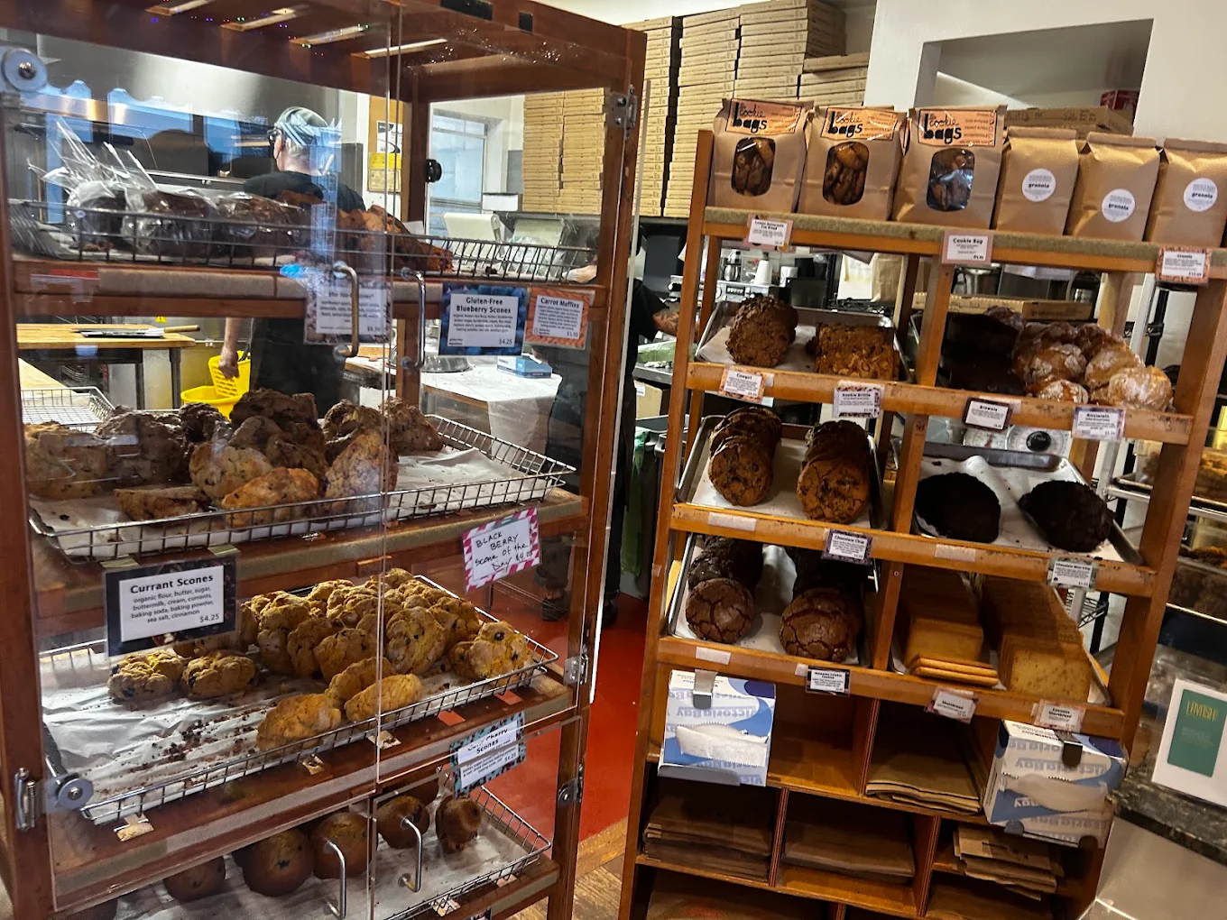 A display case and wooden shelving unit filled with various baked goods, including scones, muffins, and bags of granola, each with small price tags.