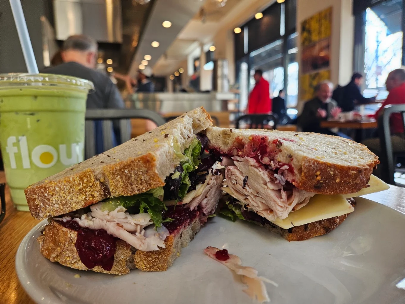 A turkey and cranberry sandwich on thick sourdough bread sits on a plate next to a green iced drink from Flour bakery.