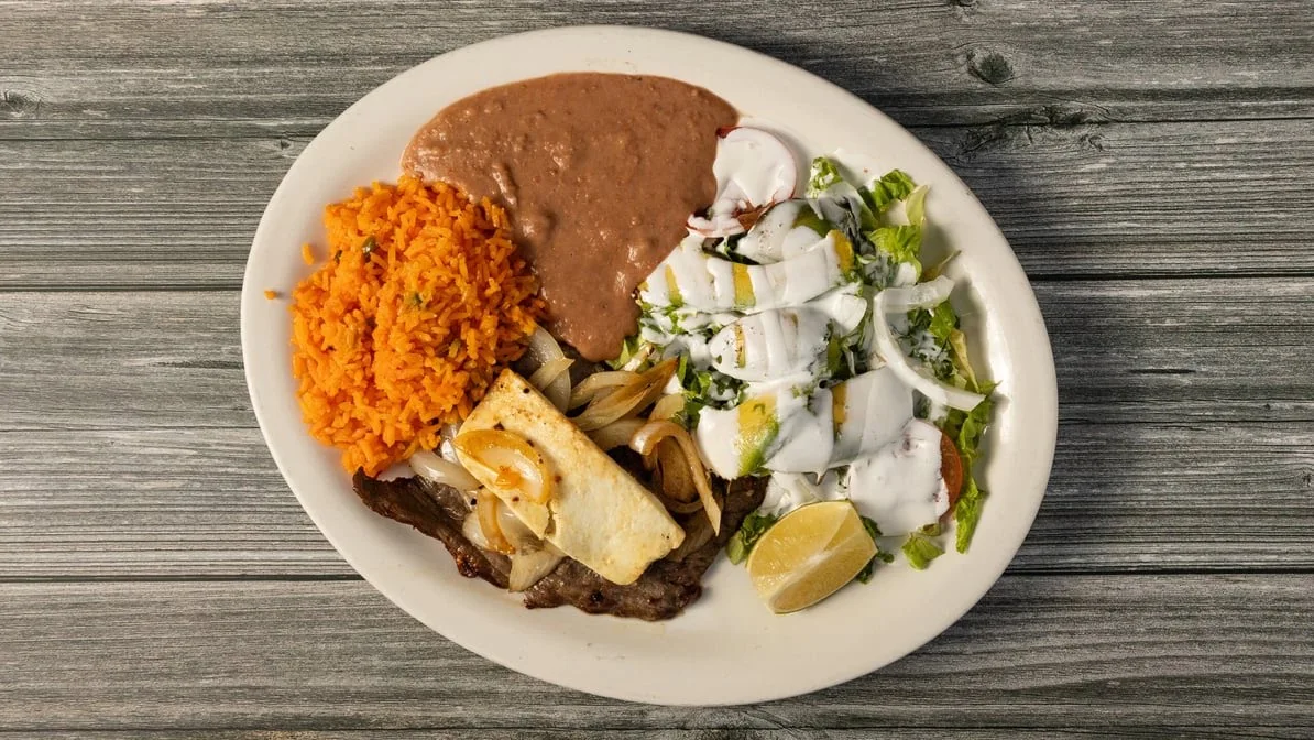 A plate of Mexican food featuring carne asada topped with grilled onions and cheese, served with orange rice, refried beans, and a side salad with dressing and a lime wedge.