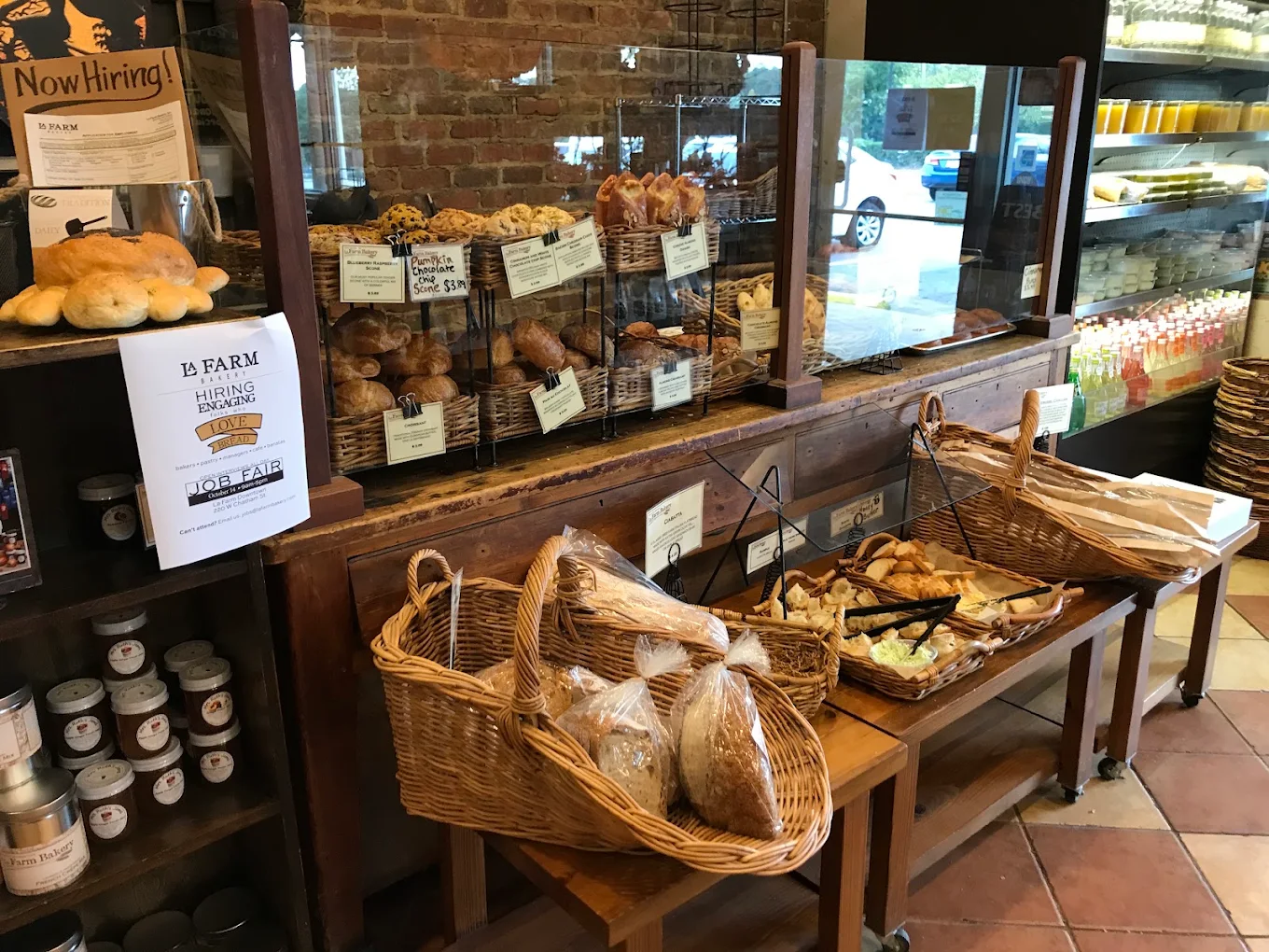 Bakery interior with a counter displaying assorted breads and pastries in wicker baskets and behind glass, with “Hiring” signs posted on the left.