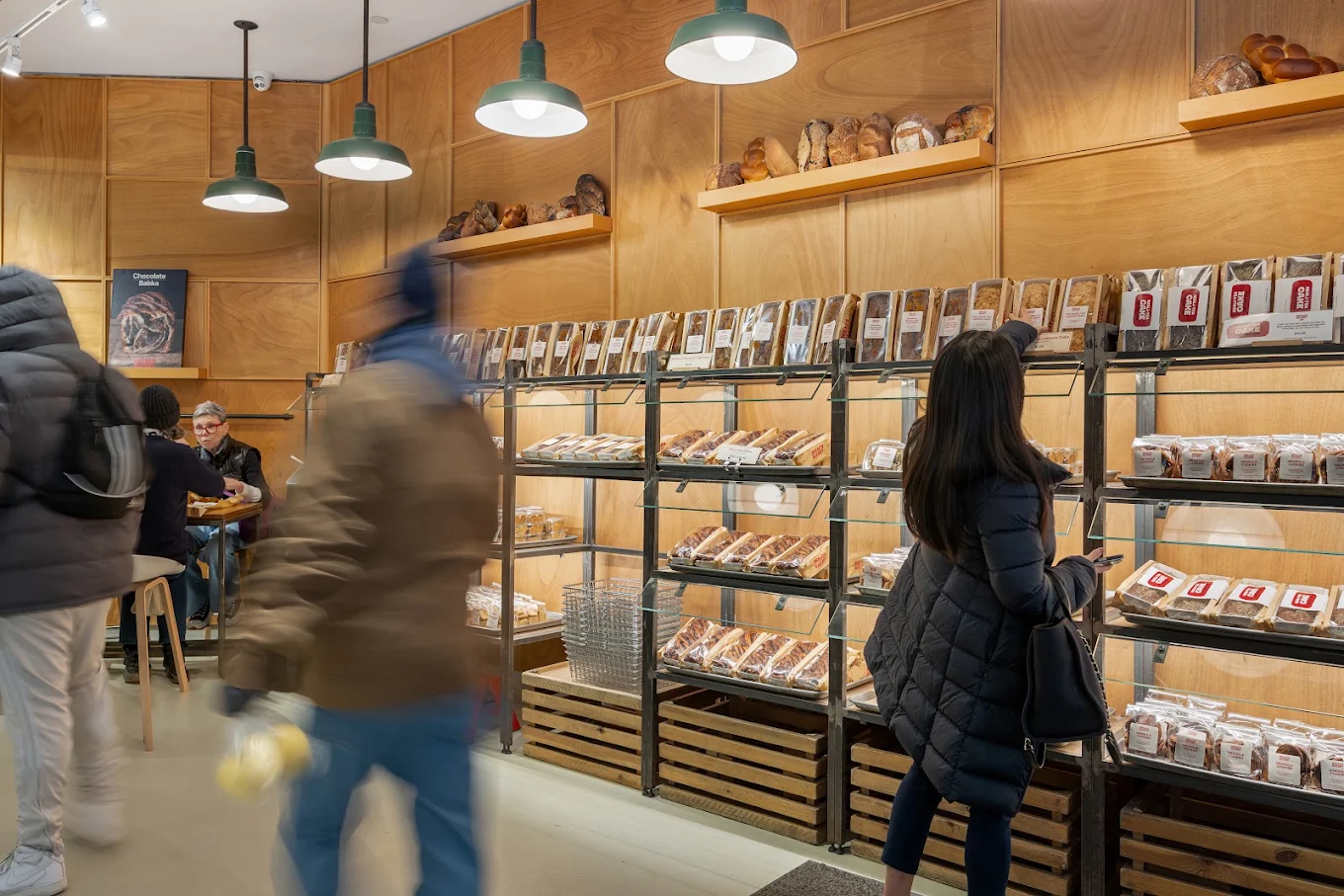 Customers browse shelves of packaged baked goods in a modern bakery with wood-paneled walls.