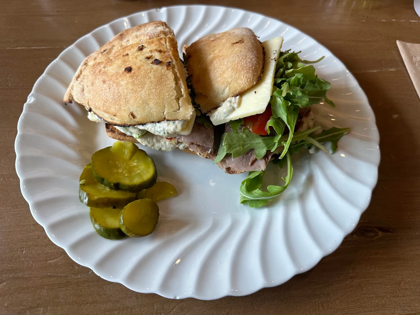 A cut panini on ciabatta bread with meat, cheese, and arugula, served with a side of sliced pickles on a white plate.