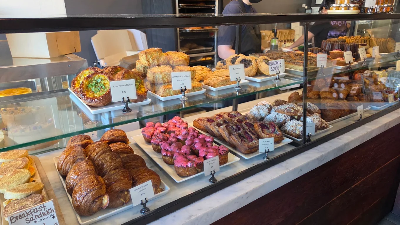 A bakery display case filled with rows of pastries, including croissants, fruit danishes, pink-glazed treats, and biscuits, each labeled with its name and price.