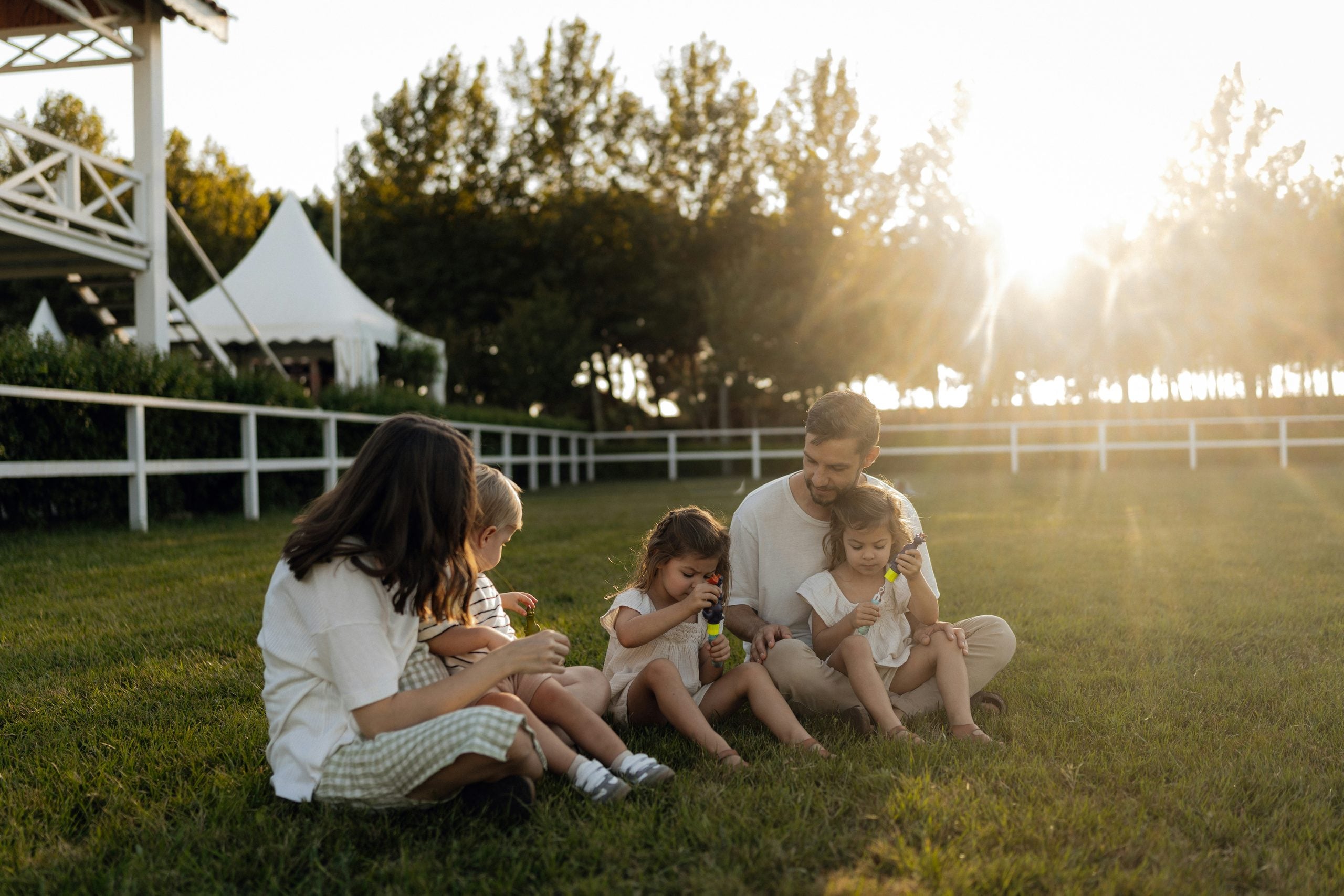 A family sits on grass blowing bubbles at sunset. Two adults and three children are together, surrounded by trees and a white fence, with sunlight shining in the background.