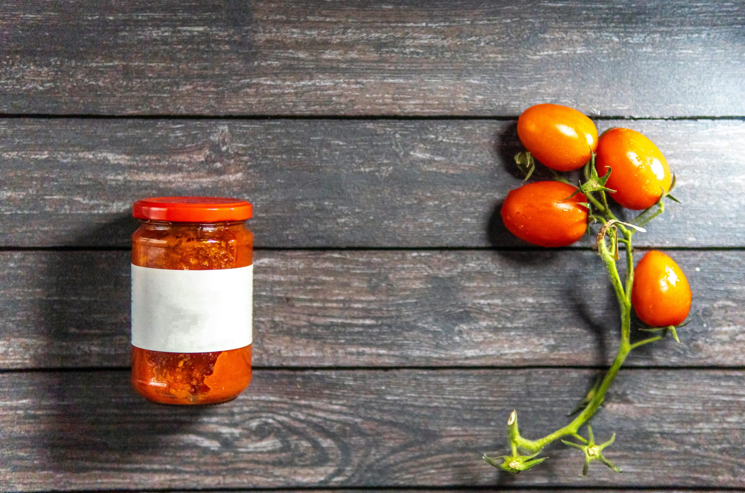 A jar with a red lid and blank label sits on a wooden surface next to a vine with five ripe tomatoes.