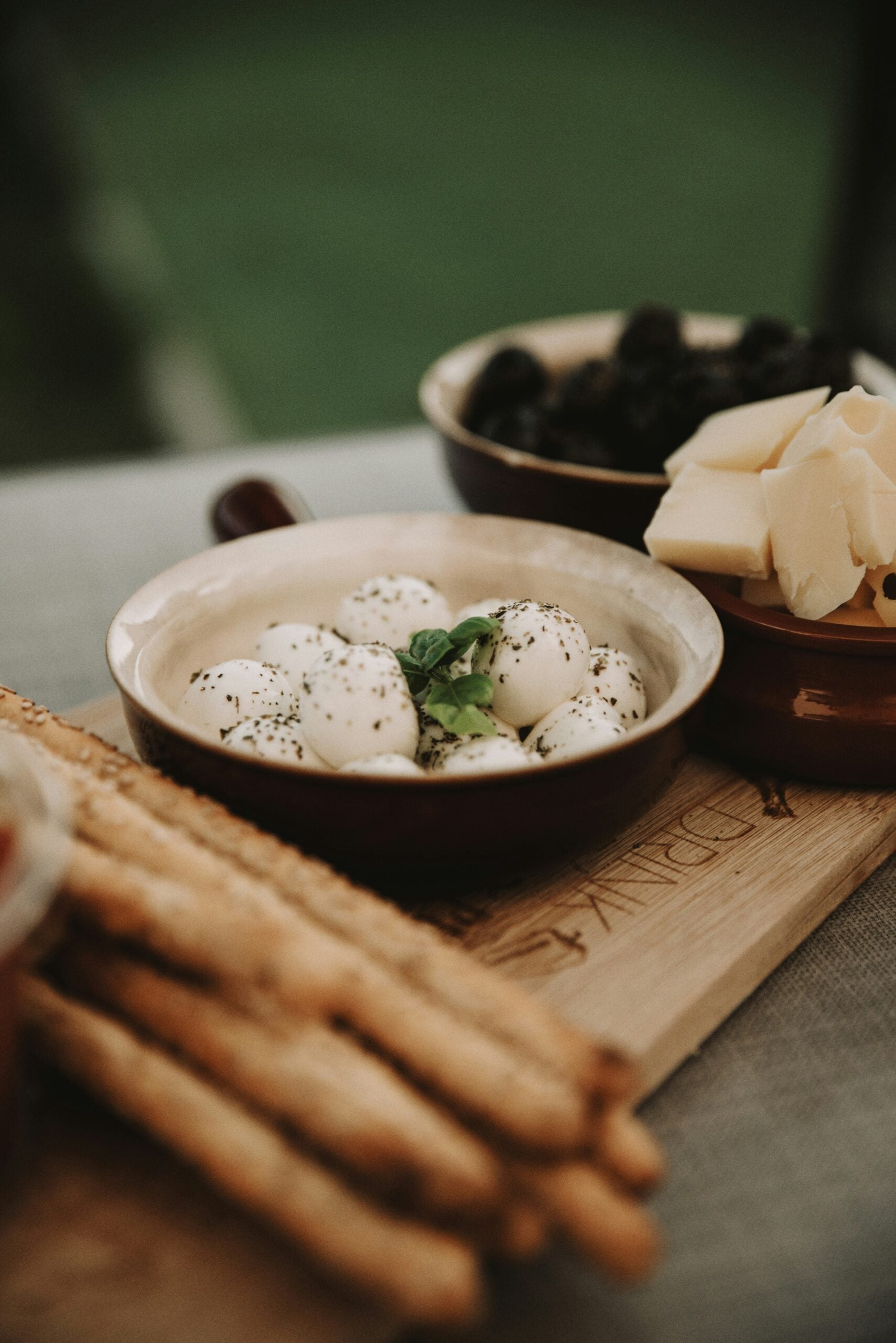 Small mozzarella balls seasoned with herbs and topped with basil in a bowl, alongside breadsticks, olives, and cheese on a wooden board.