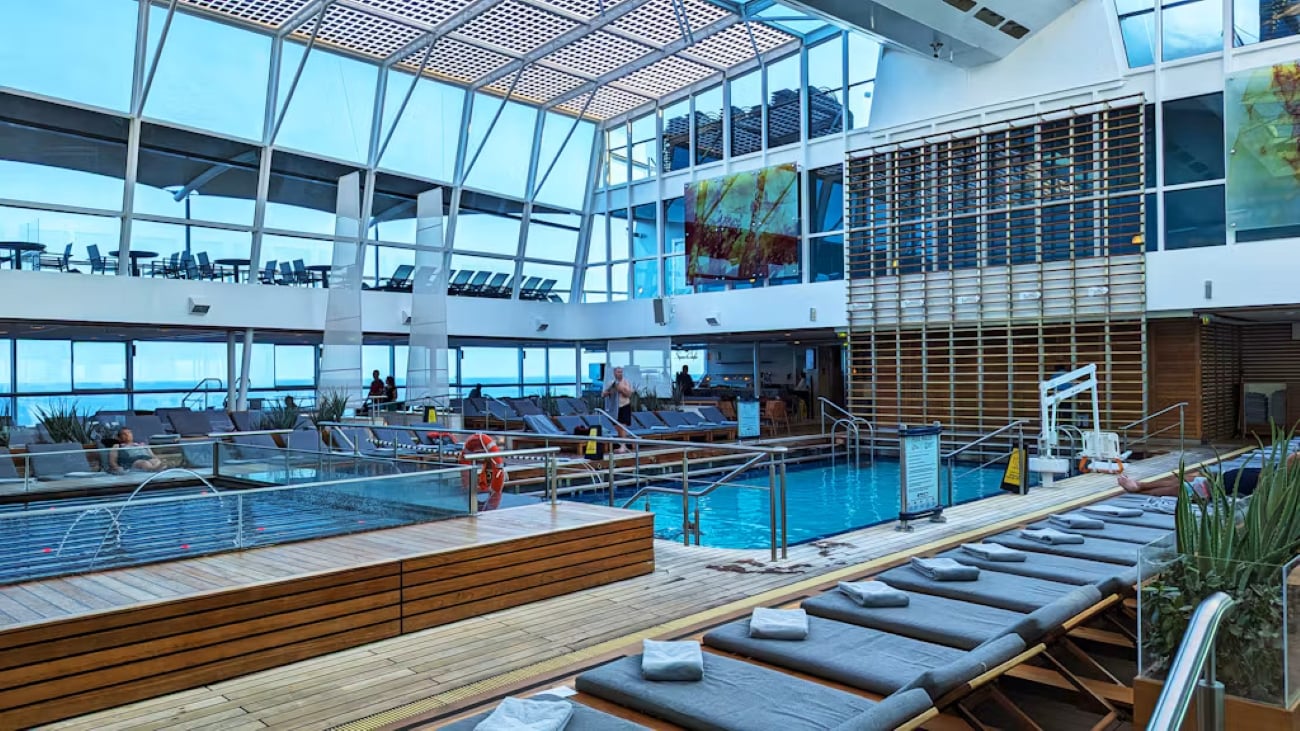 Cruise ship pools: An indoor cruise ship pool area with lounge chairs, a glass ceiling, and a wooden deck.