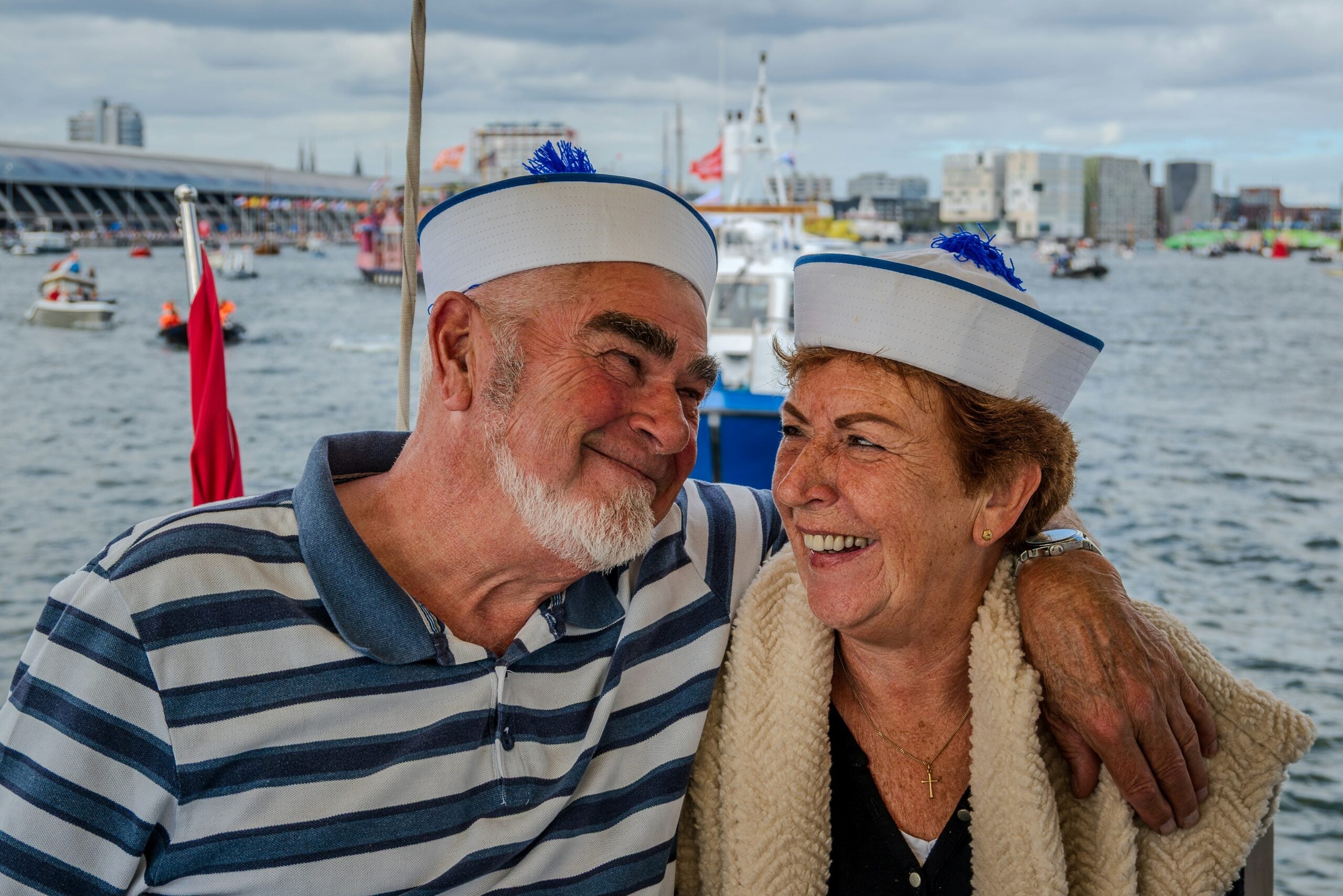 An older man and woman wearing sailor hats smile at each other on a boat, with the man’s arm around the woman. Water, boats, and city buildings are visible in the background under a cloudy sky.