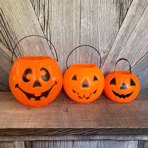Three graduated orange plastic jack-o’-lantern trick-or-treat buckets with black handles arranged in a row.